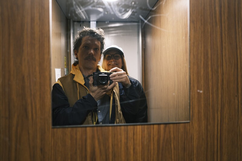 Mirror selfie of two people in a wood-panelled elevator — man with a moustache holding a small camera, woman in a cap smiling behind him