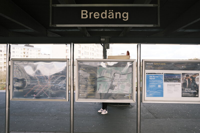 Bredäng metro station platform with the station name sign overhead and information boards below, a woman seen from behind reading them