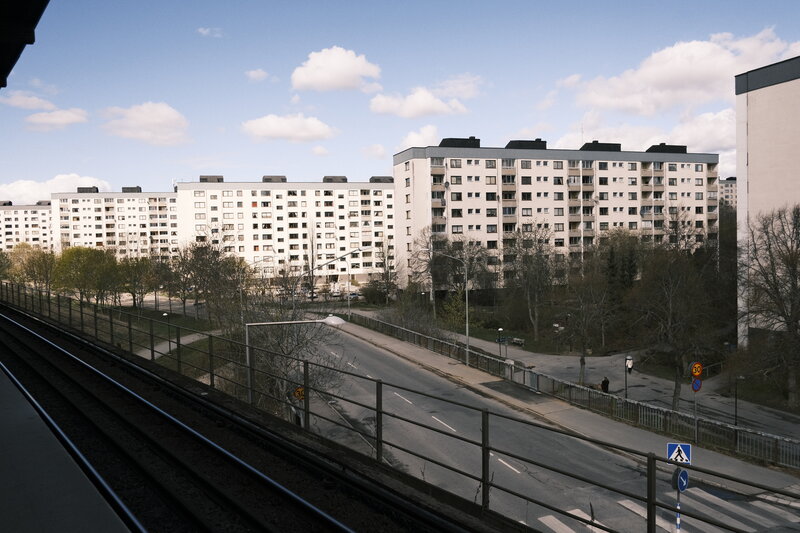 Row of white concrete apartment blocks seen across the metro tracks, bare spring trees lining the road below