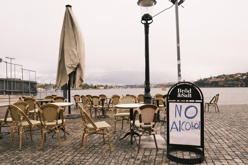 Empty outdoor terrace of Bröd & Salt on a grey day — wicker chairs around tables on cobblestone, a handwritten No Alcohol sign, waterfront in the background