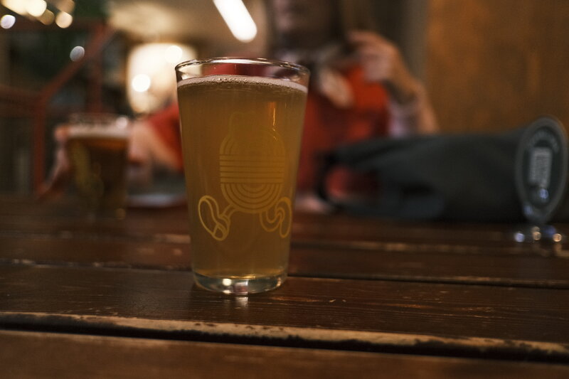 Close-up of a pint glass of pale beer on a dark wooden table, glass etched with a circular logo, warm bokeh background