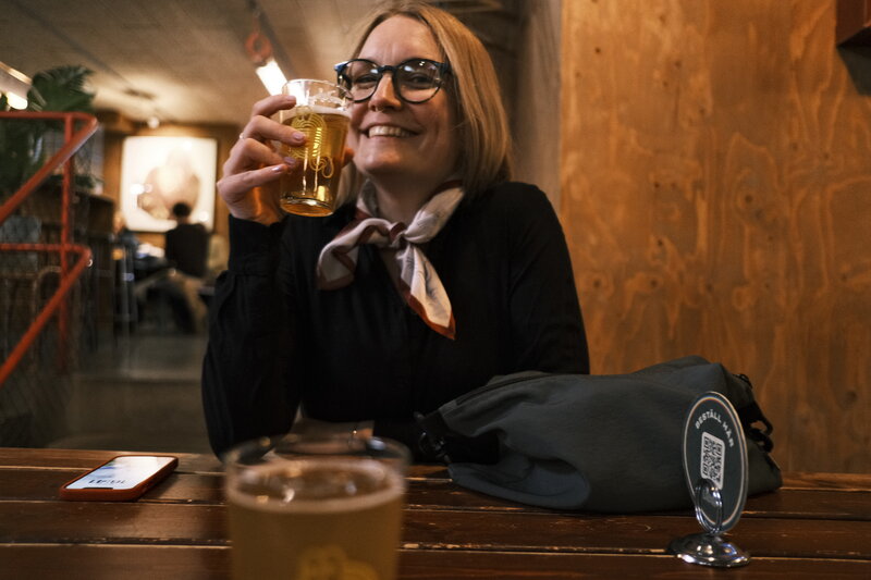 Woman with glasses laughing and raising a glass of beer at a bar table