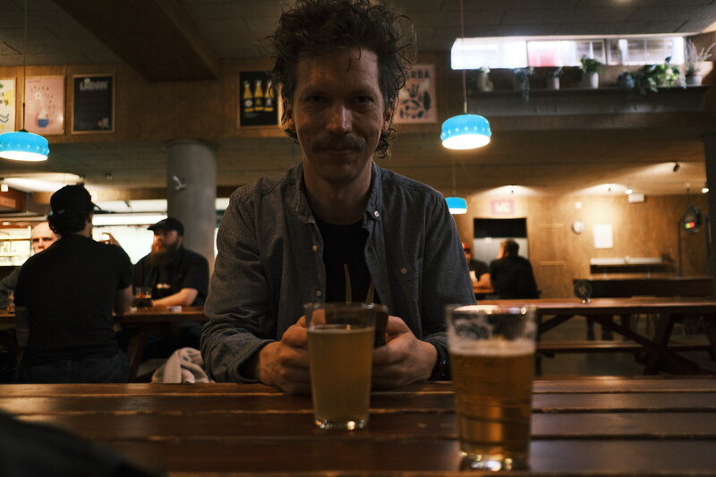 Man with curly hair and a moustache sitting at a wooden bar table holding a pint, blue pendant lights in the background