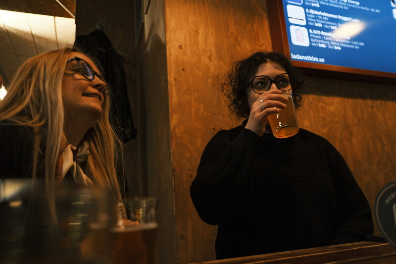 Two women at a bar counter, one drinking from a glass while the other smiles, a beer tap list on a screen behind them