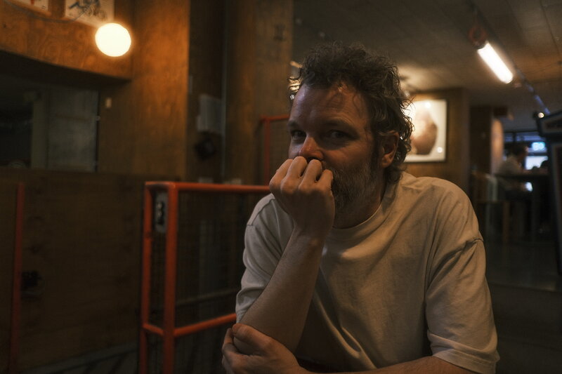 Man with curly hair resting his chin on his fist, looking off to the side in a dimly lit bar