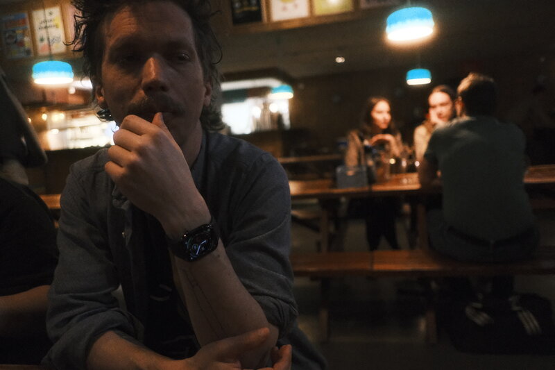 Man with curly hair and a watch leaning on his arms at a bar table, looking pensive, other patrons blurred behind him