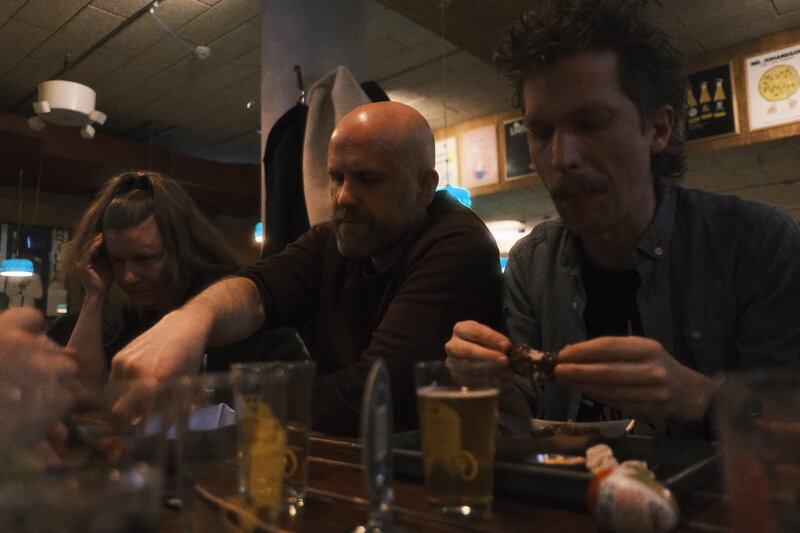 Three people hunched over a bar table — a woman and two men picking at food on a plate, beer glasses between them