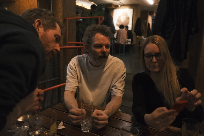 Three people around a bar table looking at something off-frame together, empty glasses in front of them