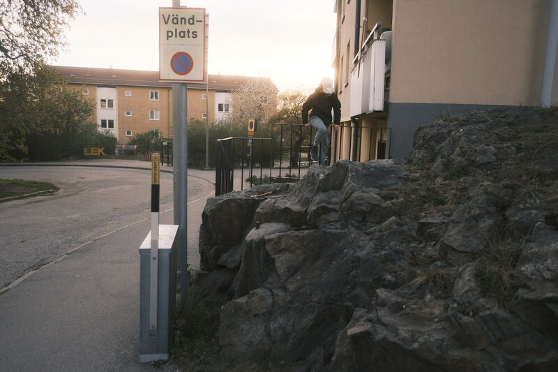 A person climbs a rocky ledge next to an apartment building, a "Vändplats" sign in the foreground with the setting sun behind