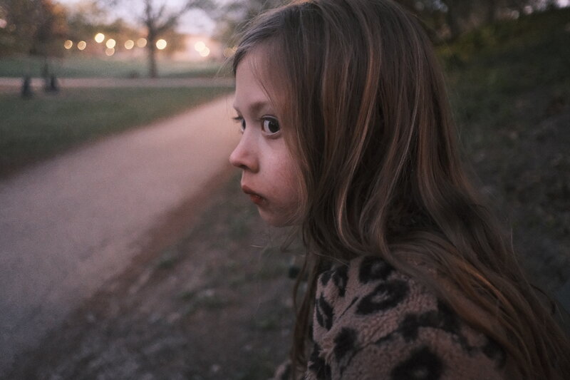 A young girl glances back over her shoulder in a park at dusk, bokeh lights visible along the path behind her